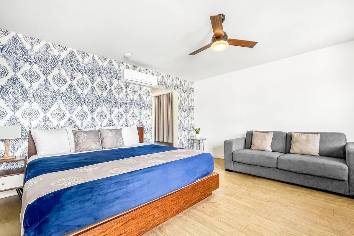 A bedroom with a bed, a grey sofa, a ceiling fan, and patterned wallpaper.