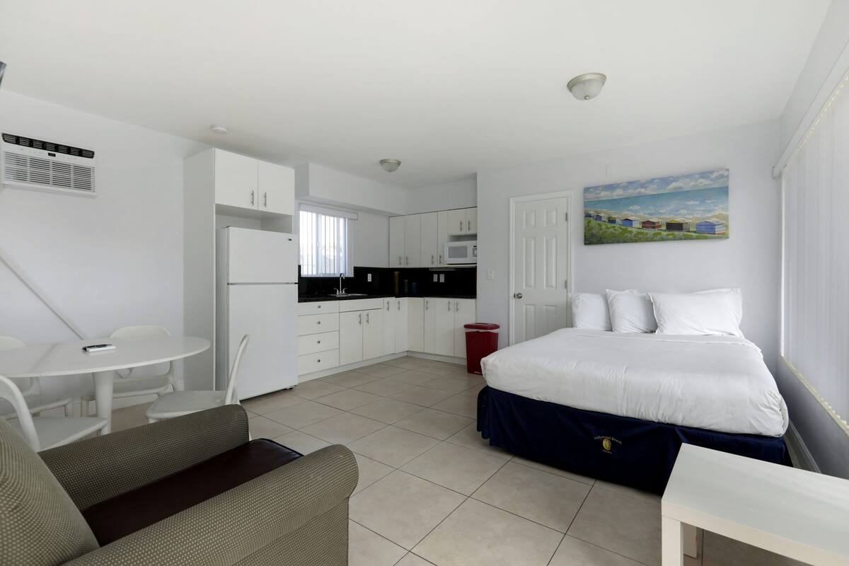 A compact hotel room with a bed, a small kitchenette, a dining area, and a painting on the wall.