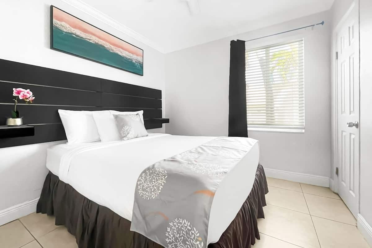 A bedroom with a bed, a nightstand, a painting, a window with blinds, and a door.