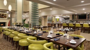 Comedor de hotel con varias mesas, sillas amarillas y una zona de bar con televisor.