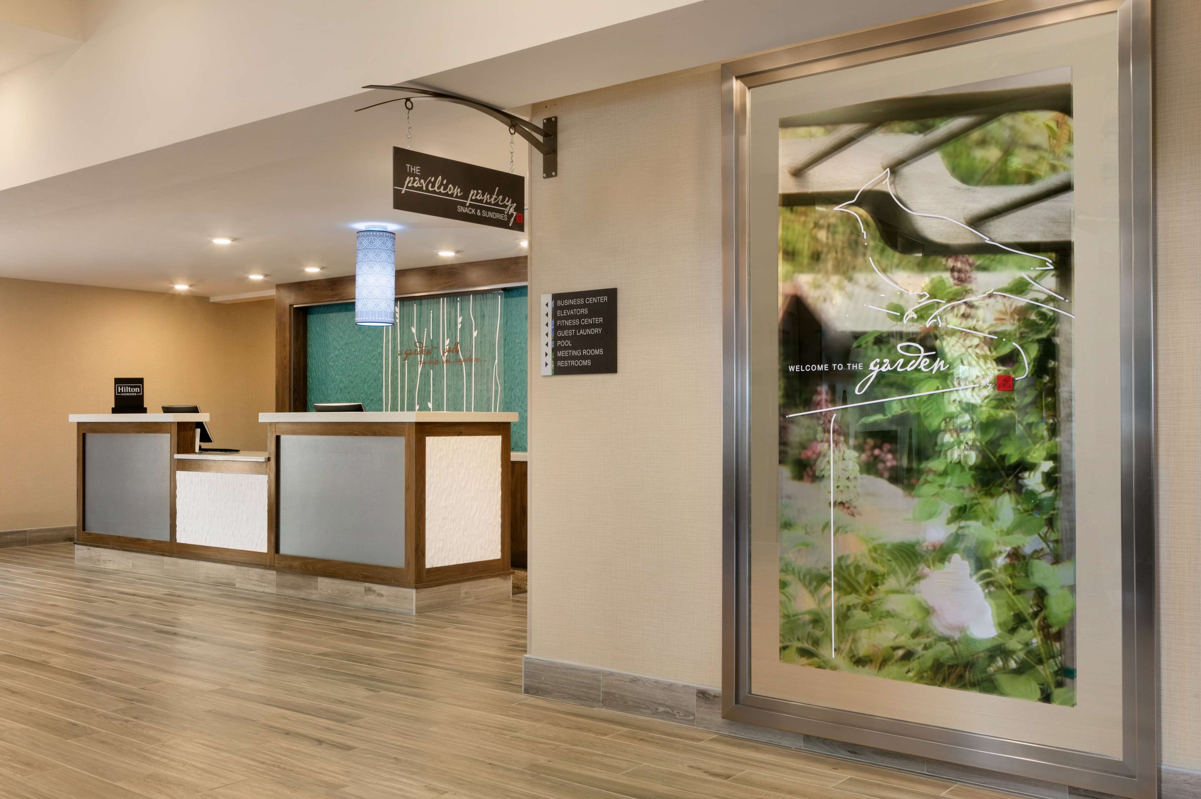 A hotel reception area with a reception desk, a business center sign, and a large poster advertising the garden.