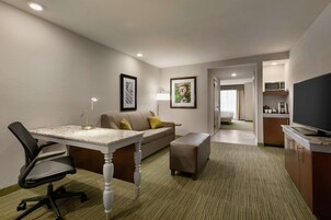 Una habitación de hotel con escritorio, silla de oficina, sofá, reposapiés y televisión.