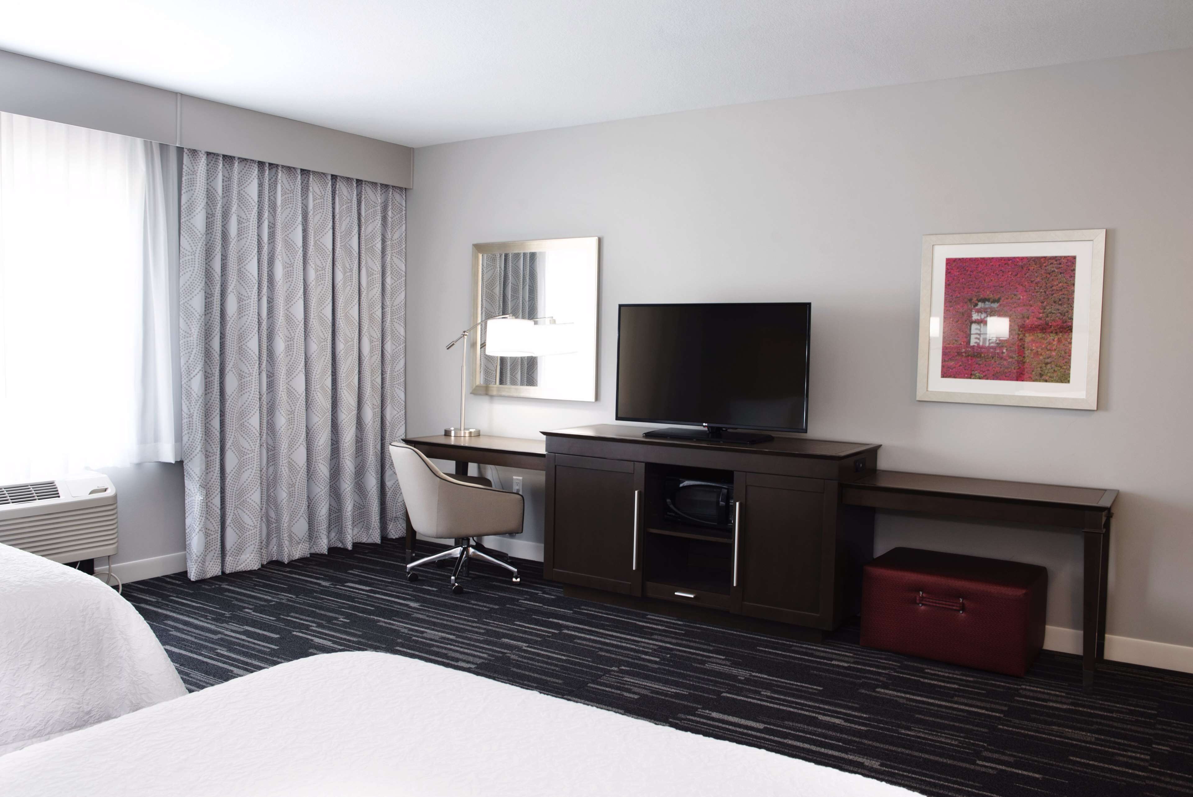 Una habitación de hotel con una cama, un escritorio con televisor, una silla, una maleta roja y una ventana con cortinas.