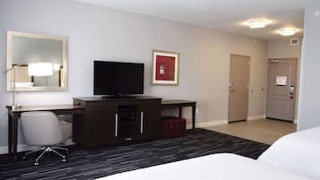 A hotel room with a bed, a desk with a computer, a TV, a mirror, a framed picture, and a door.