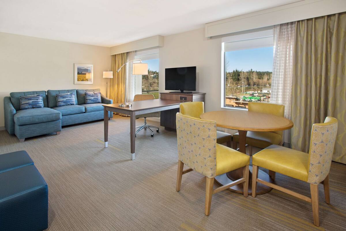 A hotel room with a sofa, a dining table with chairs, a TV, and a view of the outside.