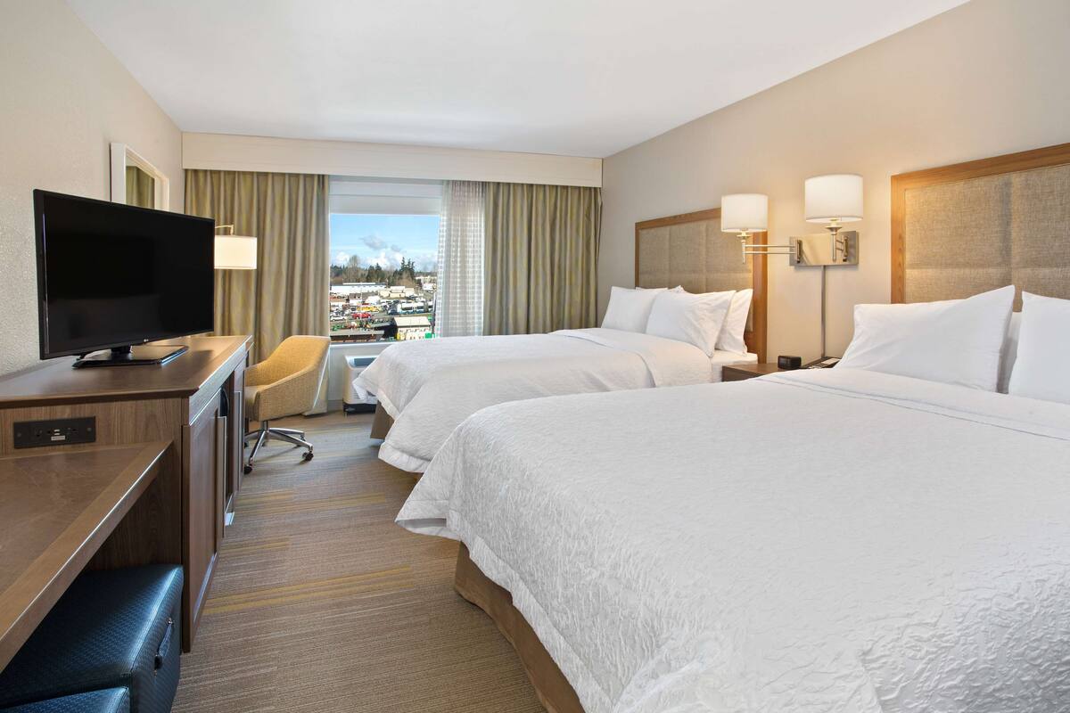 A hotel room with two beds, a desk with a television, a chair, and a view of the city.