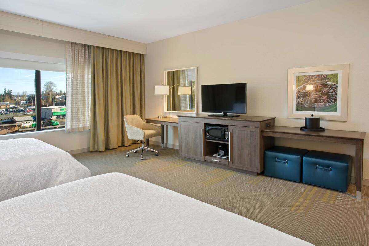 A hotel room with two beds, a desk with a TV, a chair, a desk lamp, and a window with curtains.