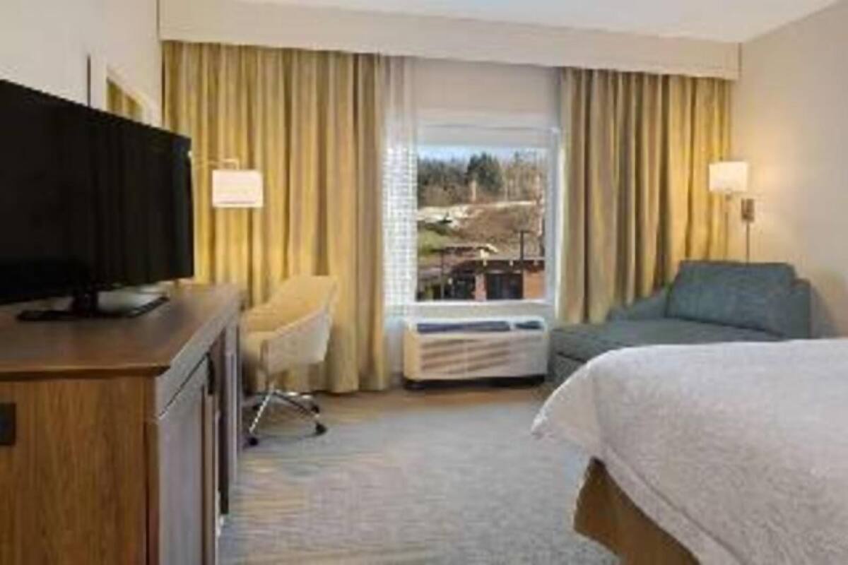 A hotel room with a bed, a desk with a television, a chair, a window with curtains, and an air conditioning unit.
