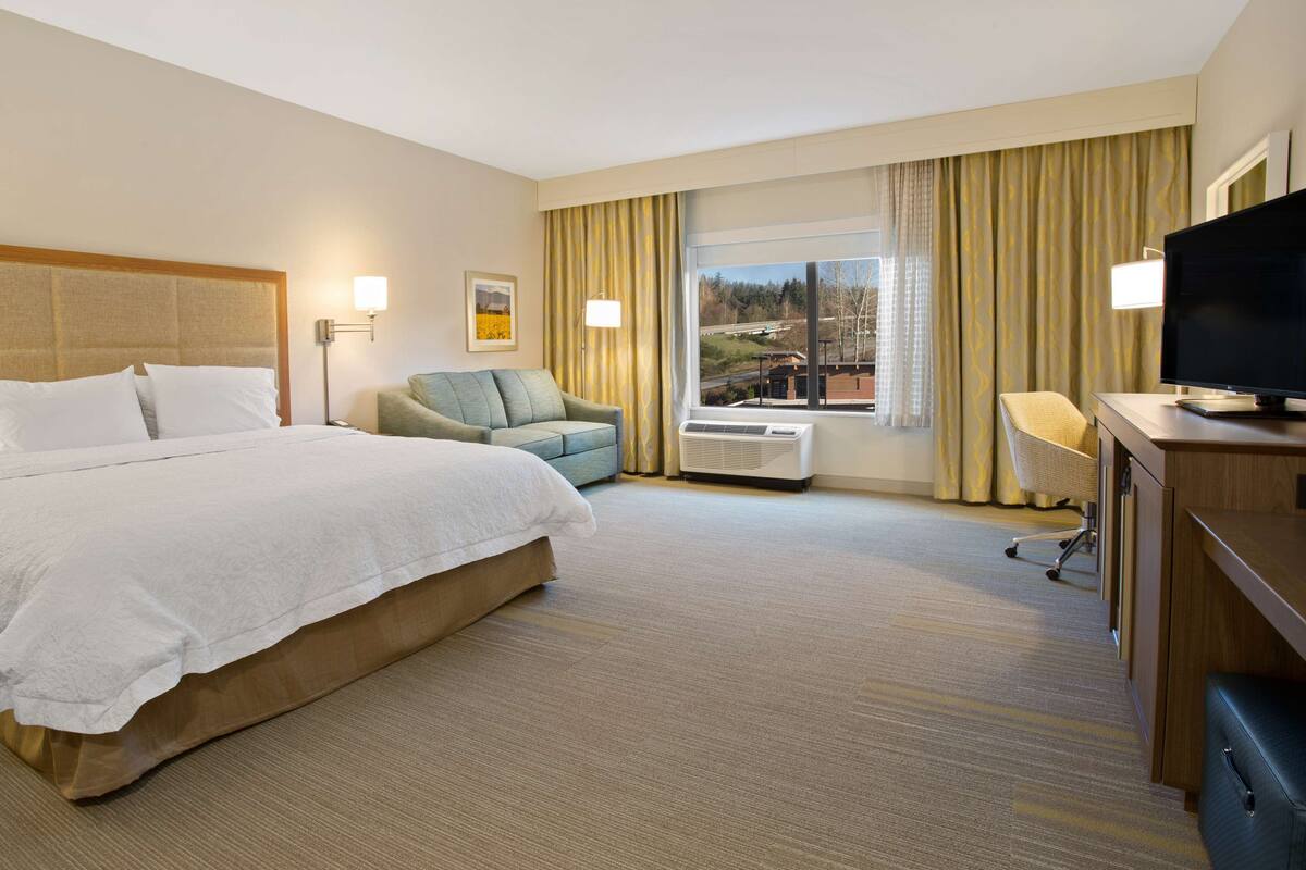 A hotel room with a large bed, a desk with a television, a chair, and a window with curtains.