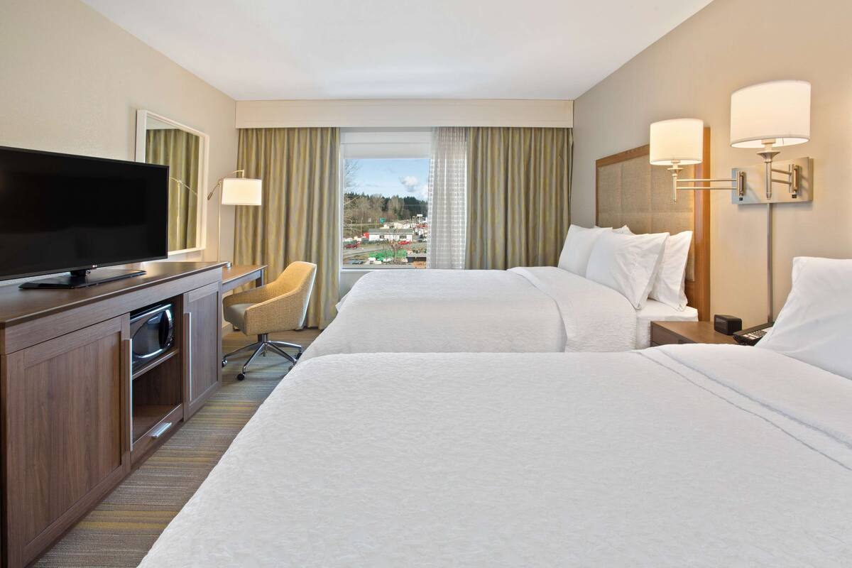 A hotel room with two beds, a TV, a desk, and a view of the city.
