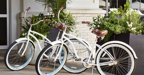 A white bicycle with blue rims parked in front of a building with a brick facade and potted plants. - The Hotel Landing (Wayzata)