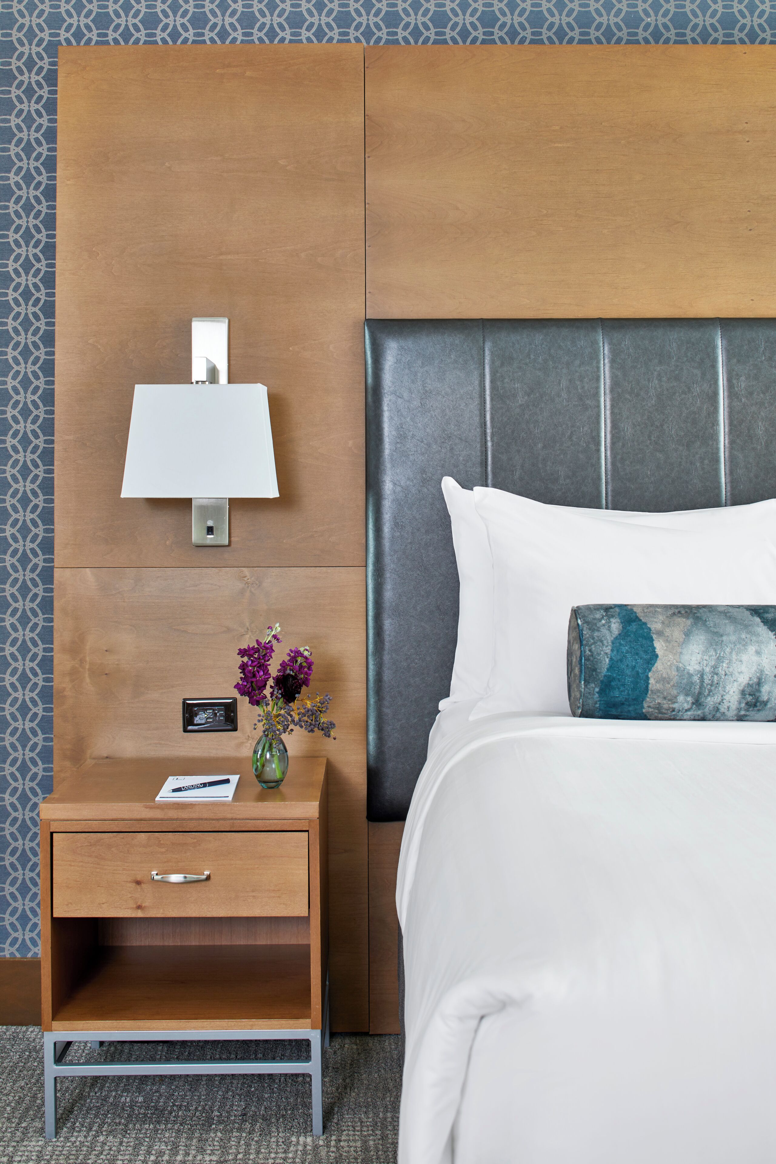 A hotel room with a wooden nightstand, a wall-mounted lamp, a vase with flowers, a bed with white bedding and a decorative pillow, and a grey headboard.