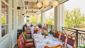 Un área de comedor al aire libre con mesas dispuestas para dos, sillas de madera y vistas a árboles y colinas.