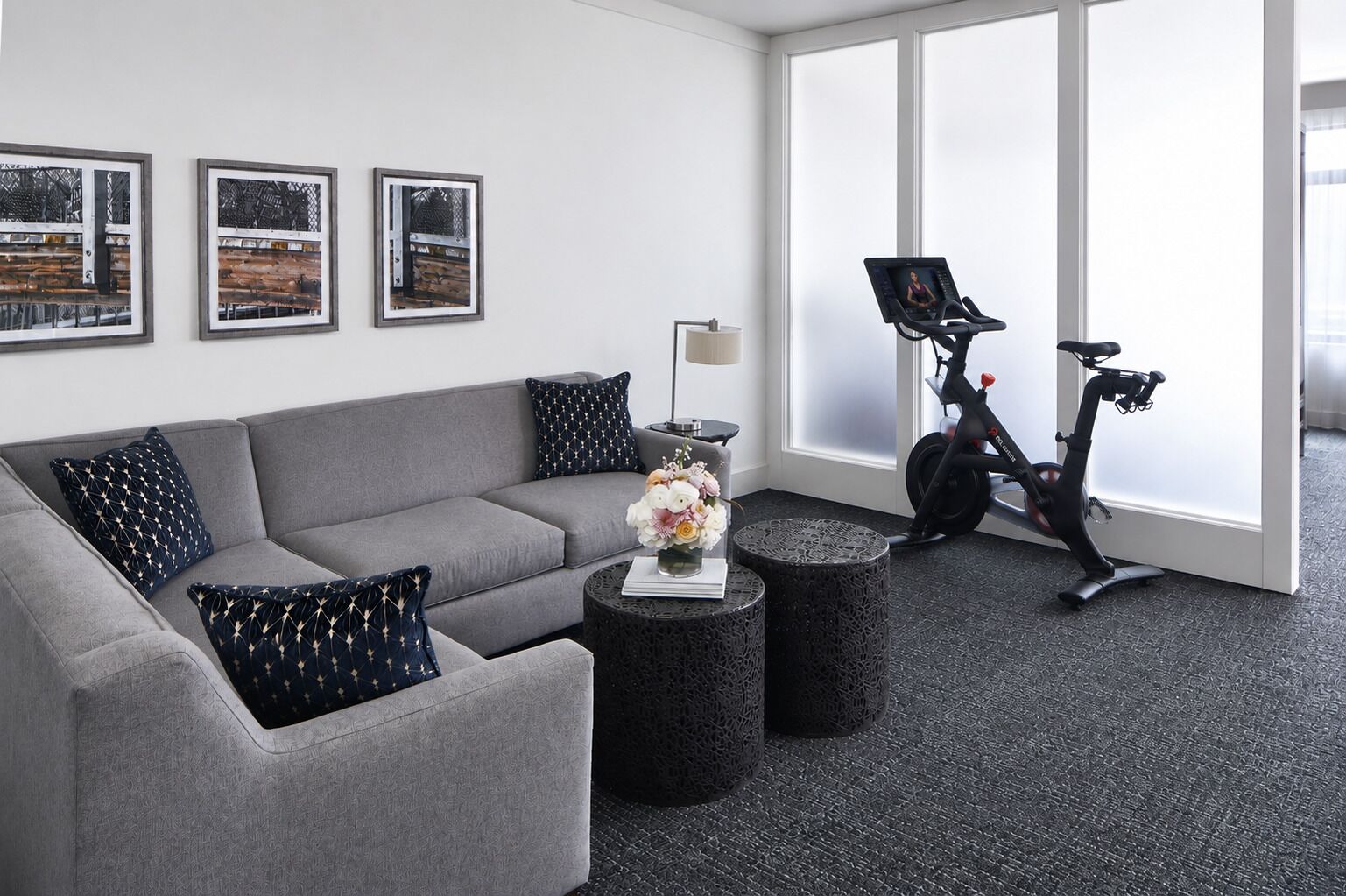 Un salon moderne avec un canapé gris, des coussins noirs à motifs, une petite table ronde ornée d’un vase de fleurs et un vélo d’appartement.