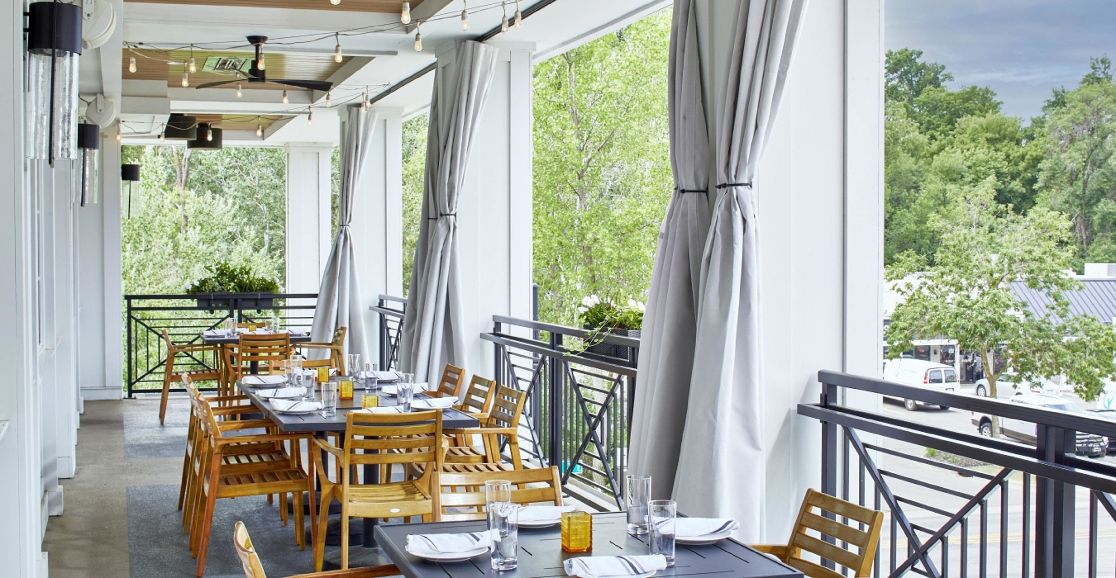 An outdoor dining area with wooden tables and chairs, large windows, and a view of trees.
