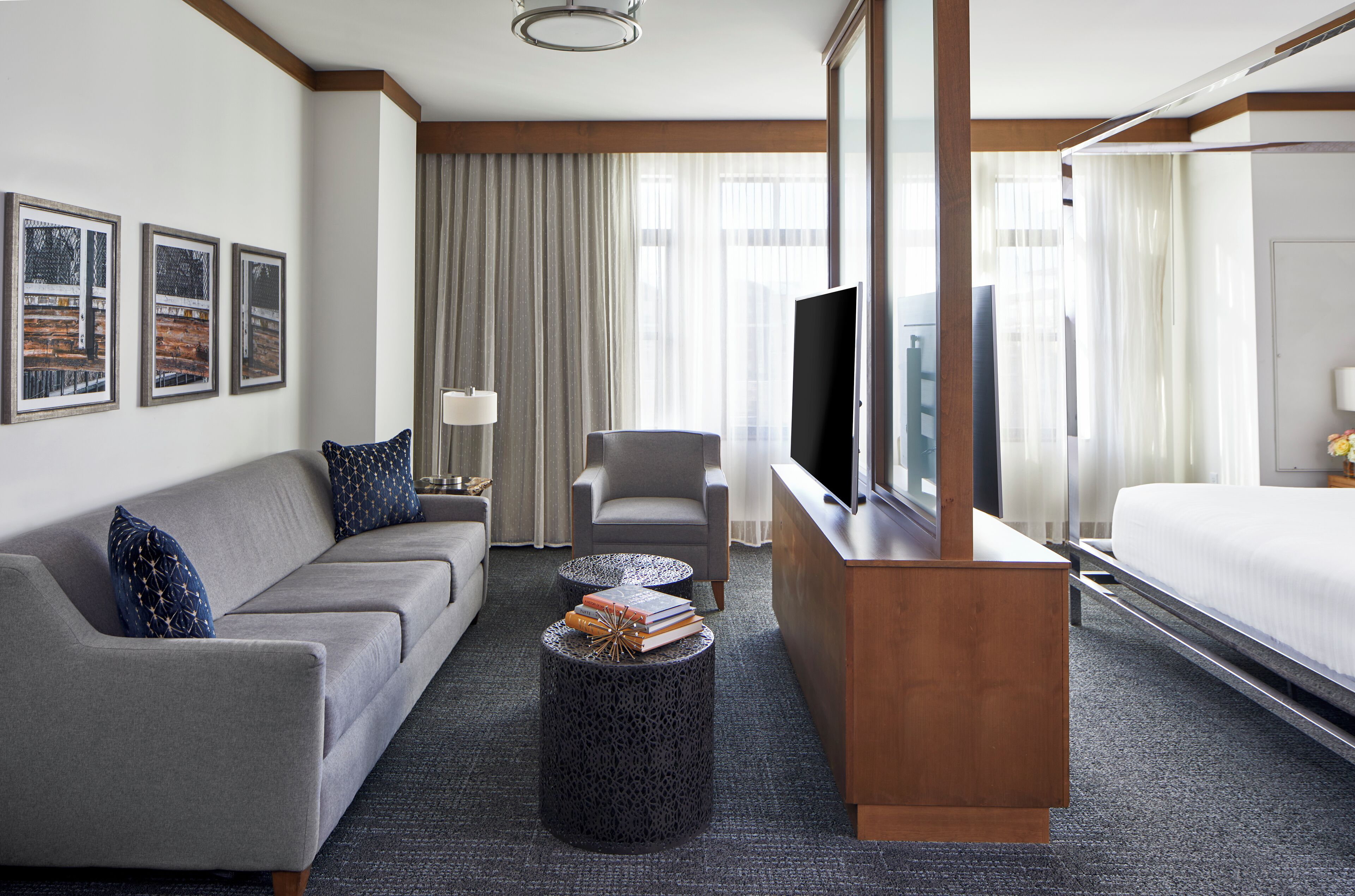 Una habitación de hotel con un sofá gris, una mesita redonda con libros, un televisor sobre un mueble de madera y una cama con edredón blanco.