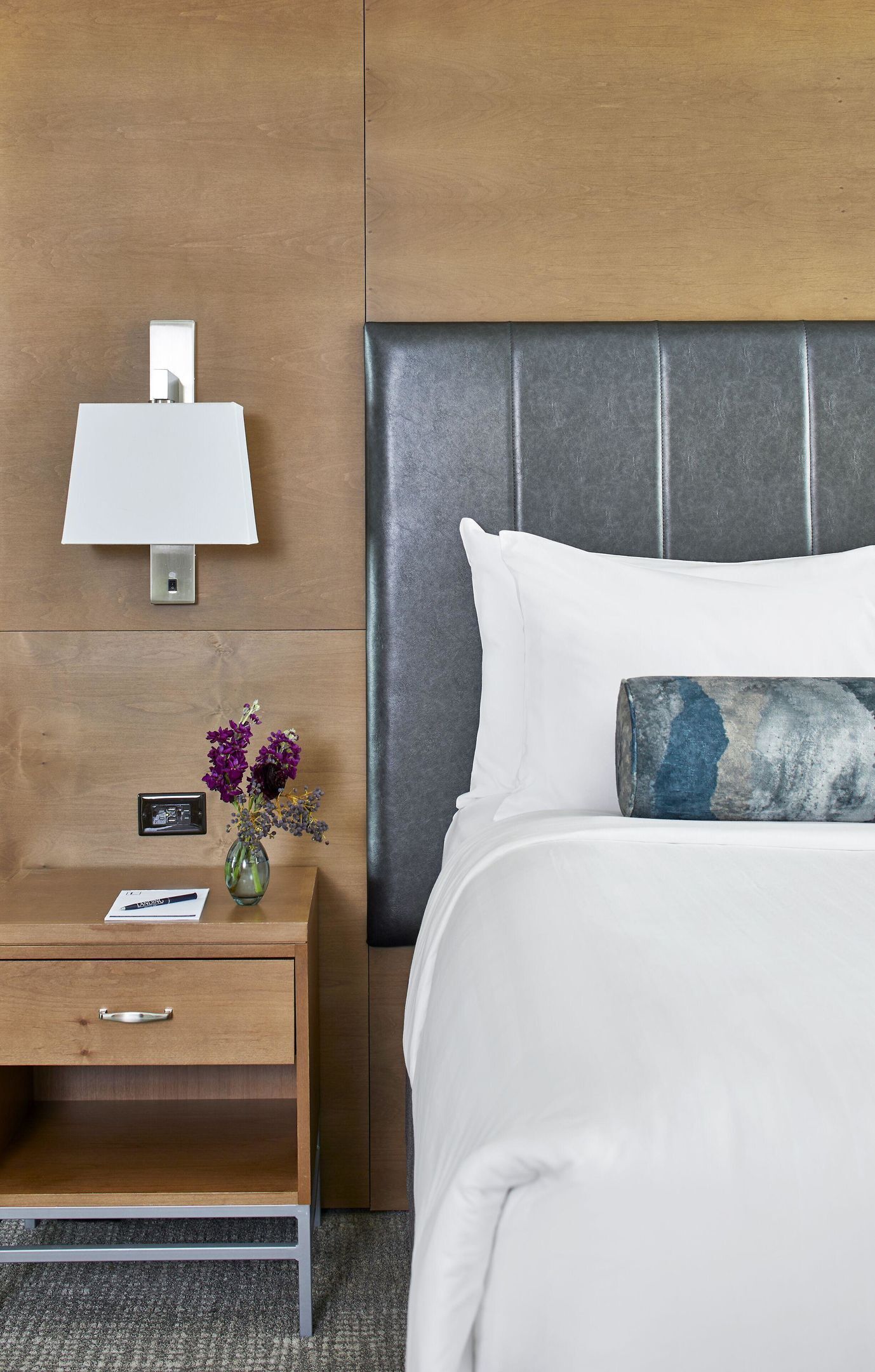 Ein Hotelzimmer mit Holzbeistelltisch, einer Lampe, einer Blumenvase, einem Bett mit weißer Bettwäsche und einem grauen Kopfteil.