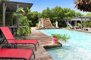 A wooden deck with red lounge chairs, a clear pool, a stone waterfall, and a shaded area with a thatched roof. - Red Palm Village (Kralendijk)