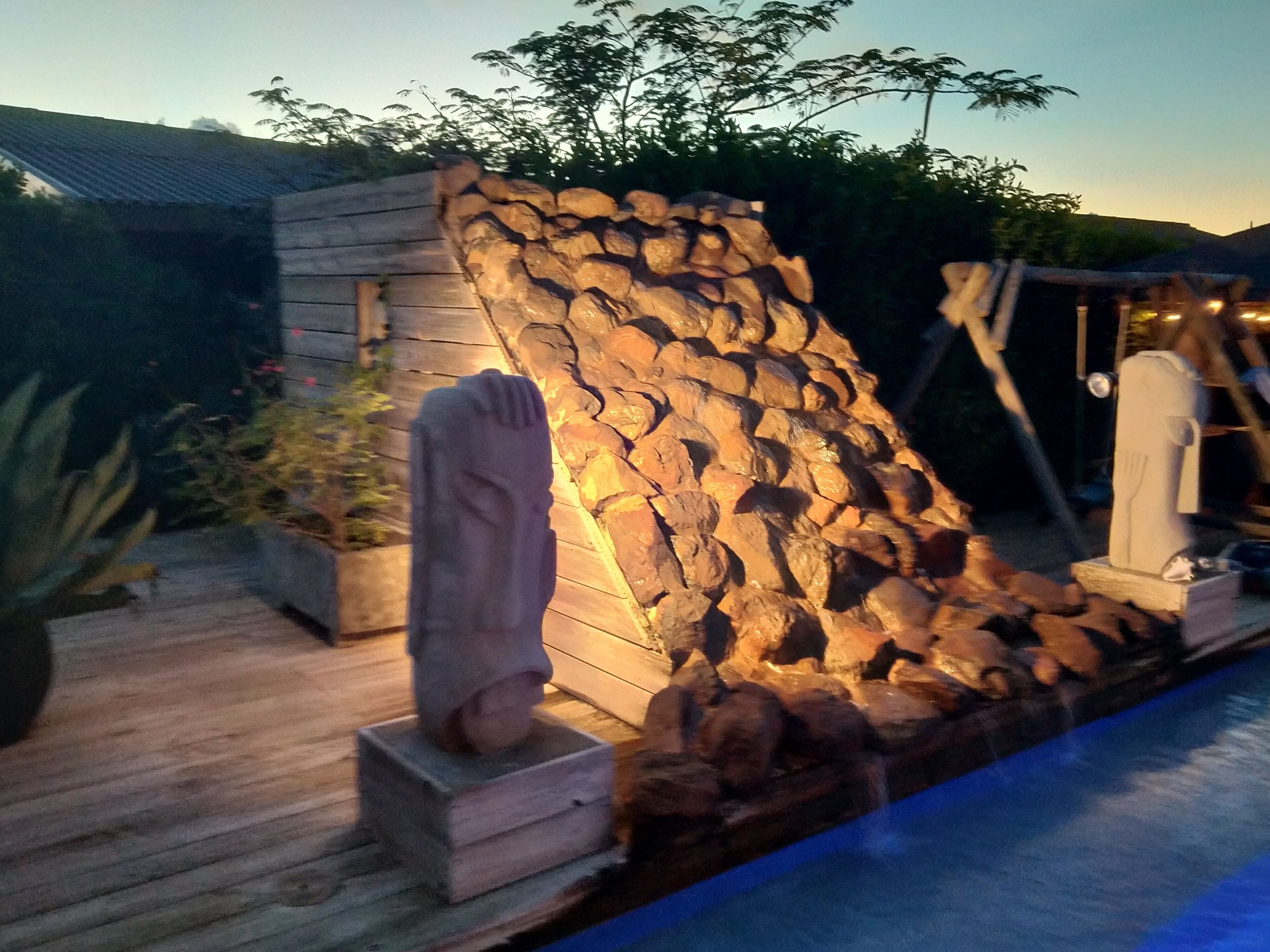 Een stenen muur met een standbeeld ervoor, een houten terras en een zwembad.