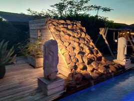 A stone wall with a statue in front, a wooden deck, and a pool.