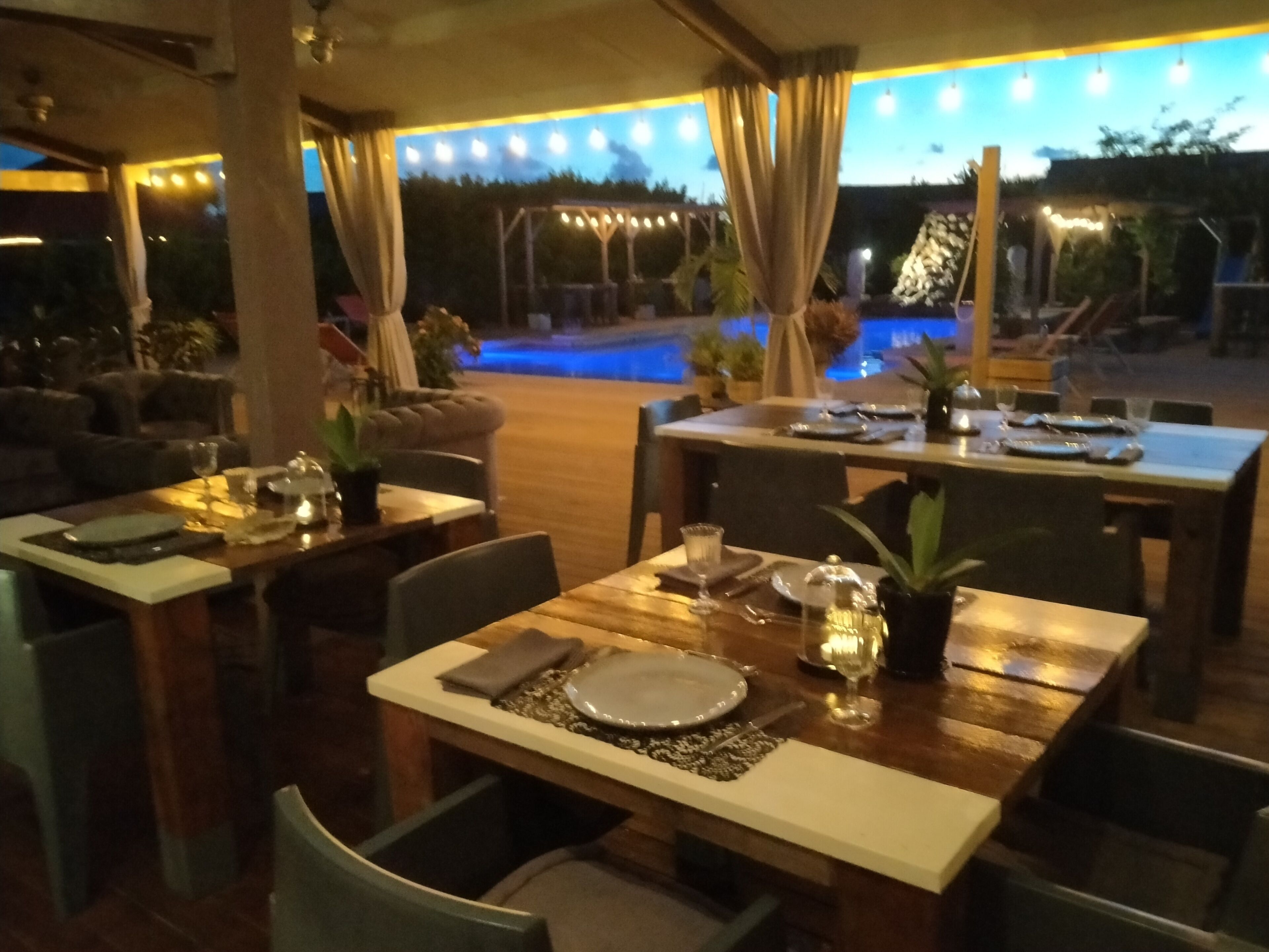 An outdoor dining area with tables set for two, featuring a pool in the background.