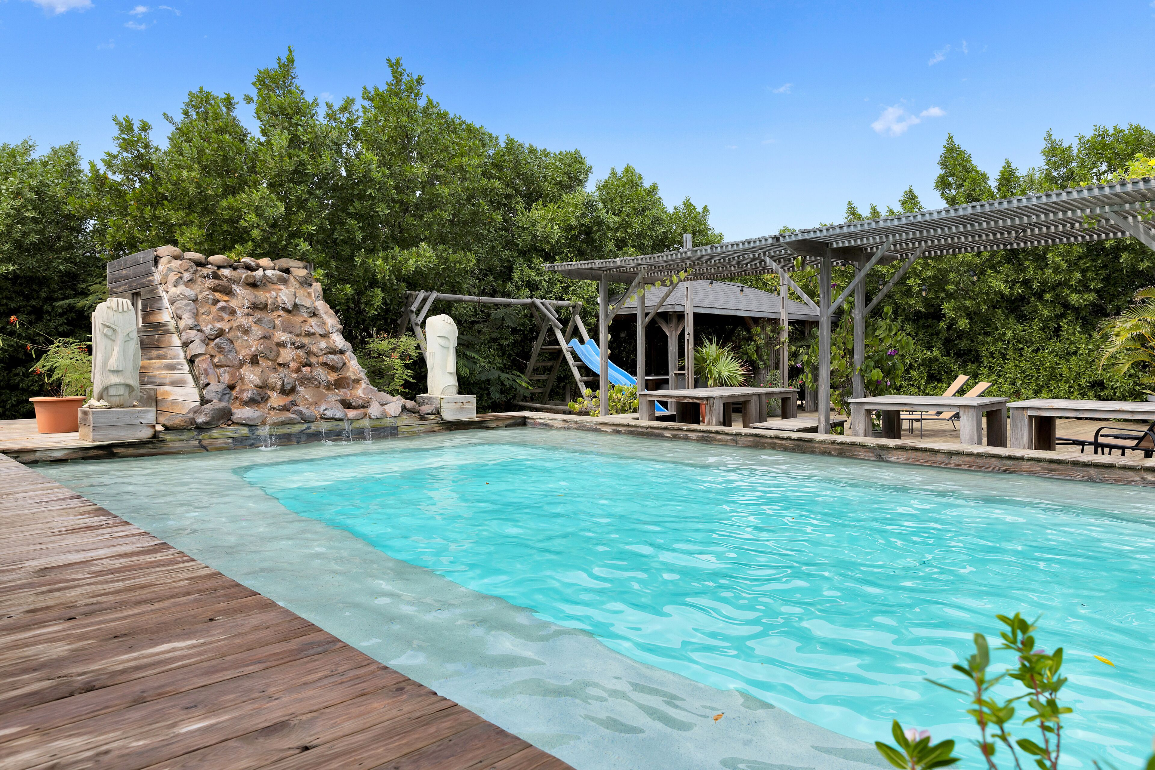Una piscina con muelle de madera, pared de roca, tobogán y un pabellón.