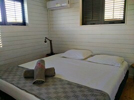 Una cama individual con ropa de cama blanca, una toalla enrollada y una manta estampada. Hay una lámpara de noche y una ventana con persianas.