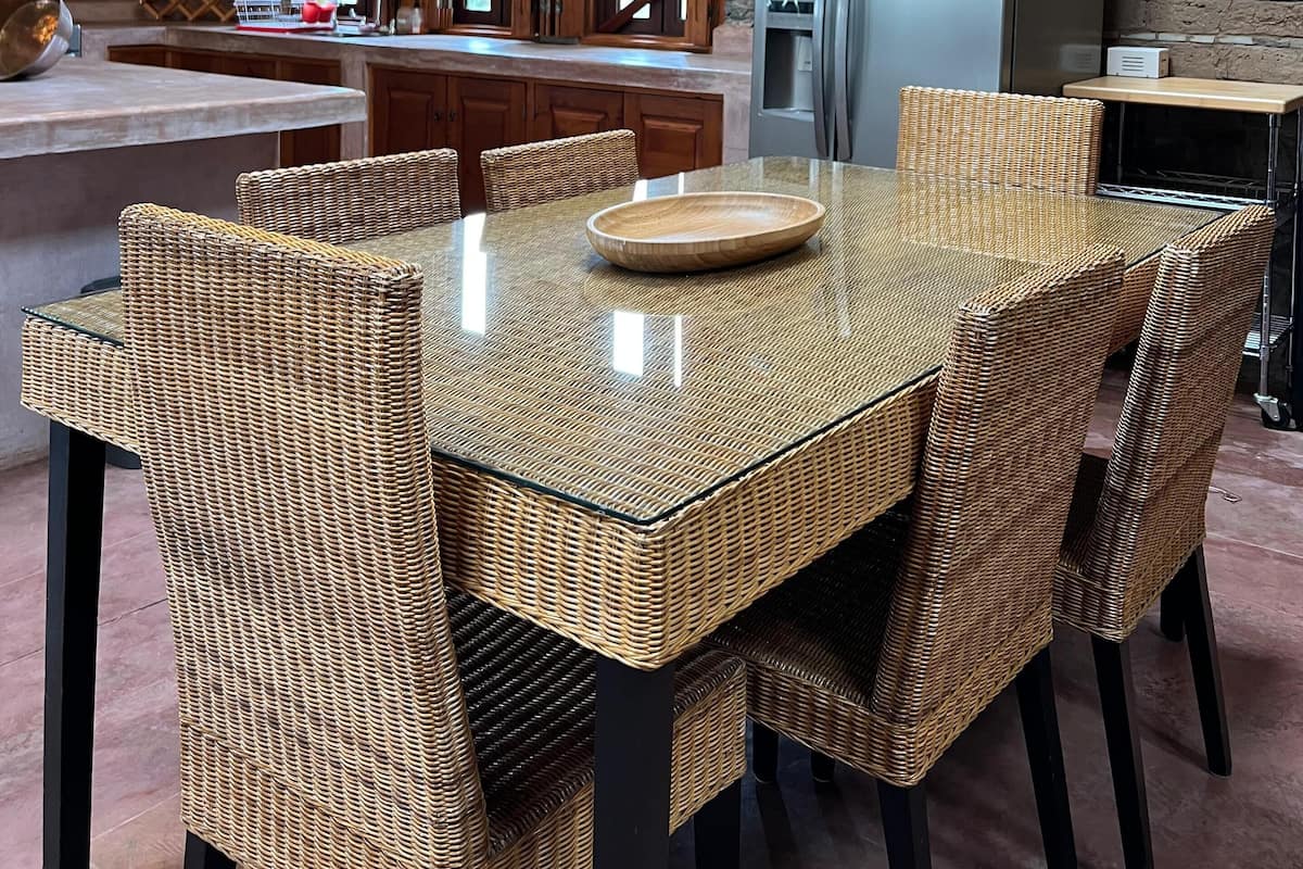Une cuisine rustique aux murs de pierre, aux poutres apparentes et dotée d’un grand îlot central agrémenté de chaises en osier.