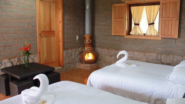 Habitación de hotel con dos camas, una pared de piedra, una puerta de madera, chimenea y ventana con contraventanas de madera.