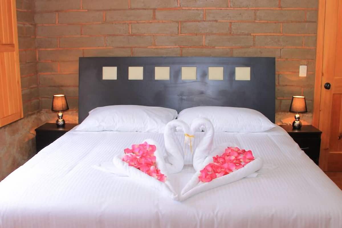 Une chambre d’hôtel avec un mur en briques, un lit recouvert de draps blancs, deux tables de chevet avec des lampes et une décoration en forme de cœur faite de serviettes et de pétales de rose.