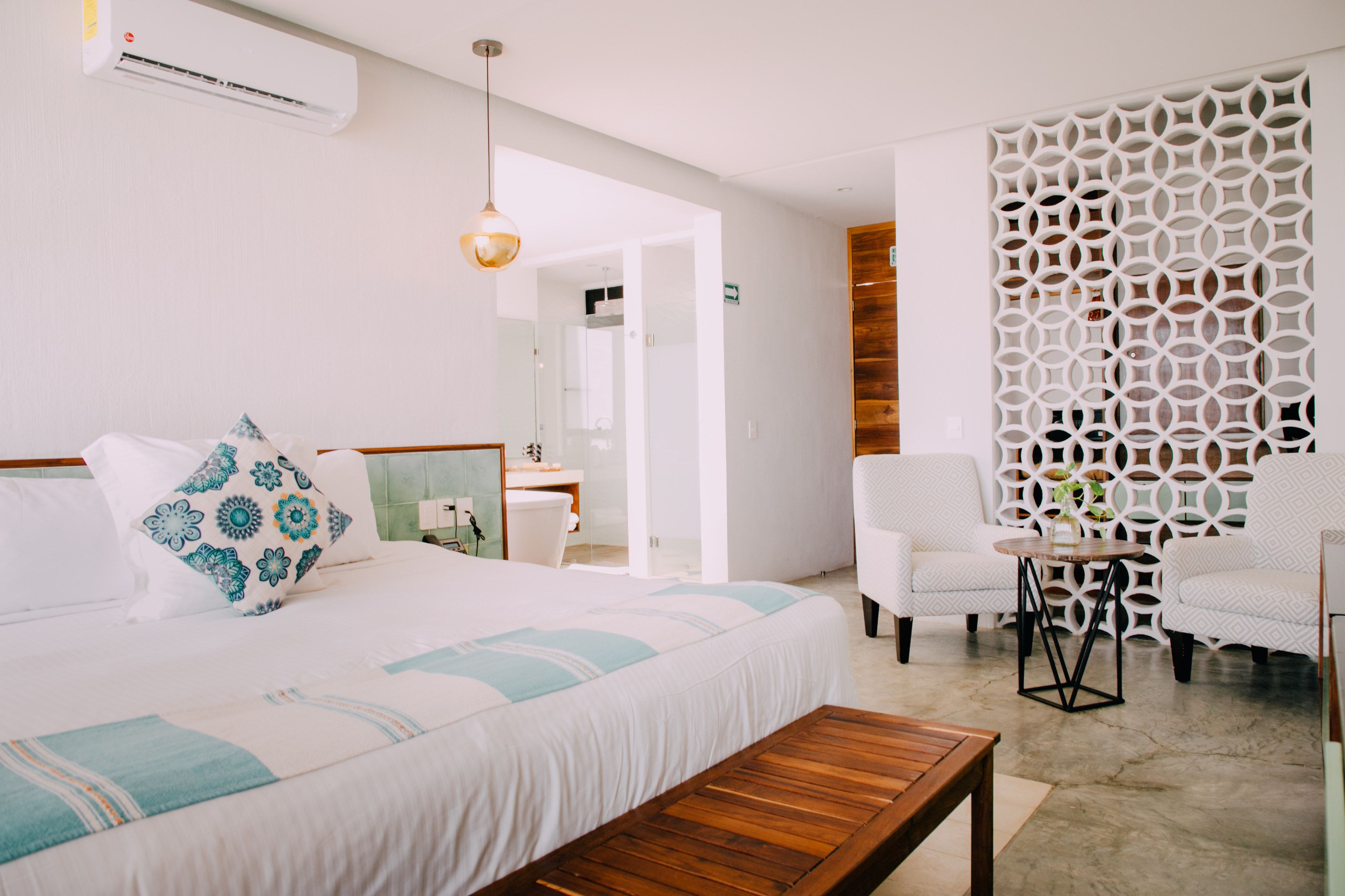 A modern hotel room with a large bed, a wooden bench, two armchairs, a small table, and a decorative white wall panel.
