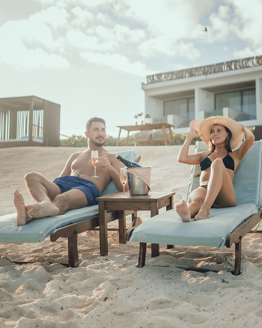 Two people relaxing on sun loungers on a sandy beach with a table between them, holding drinks and a bottle.