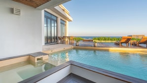 Piscina infinita no terraço com espreguiçadeiras de madeira e plantas em vasos, com vista para o céu azul e o oceano.