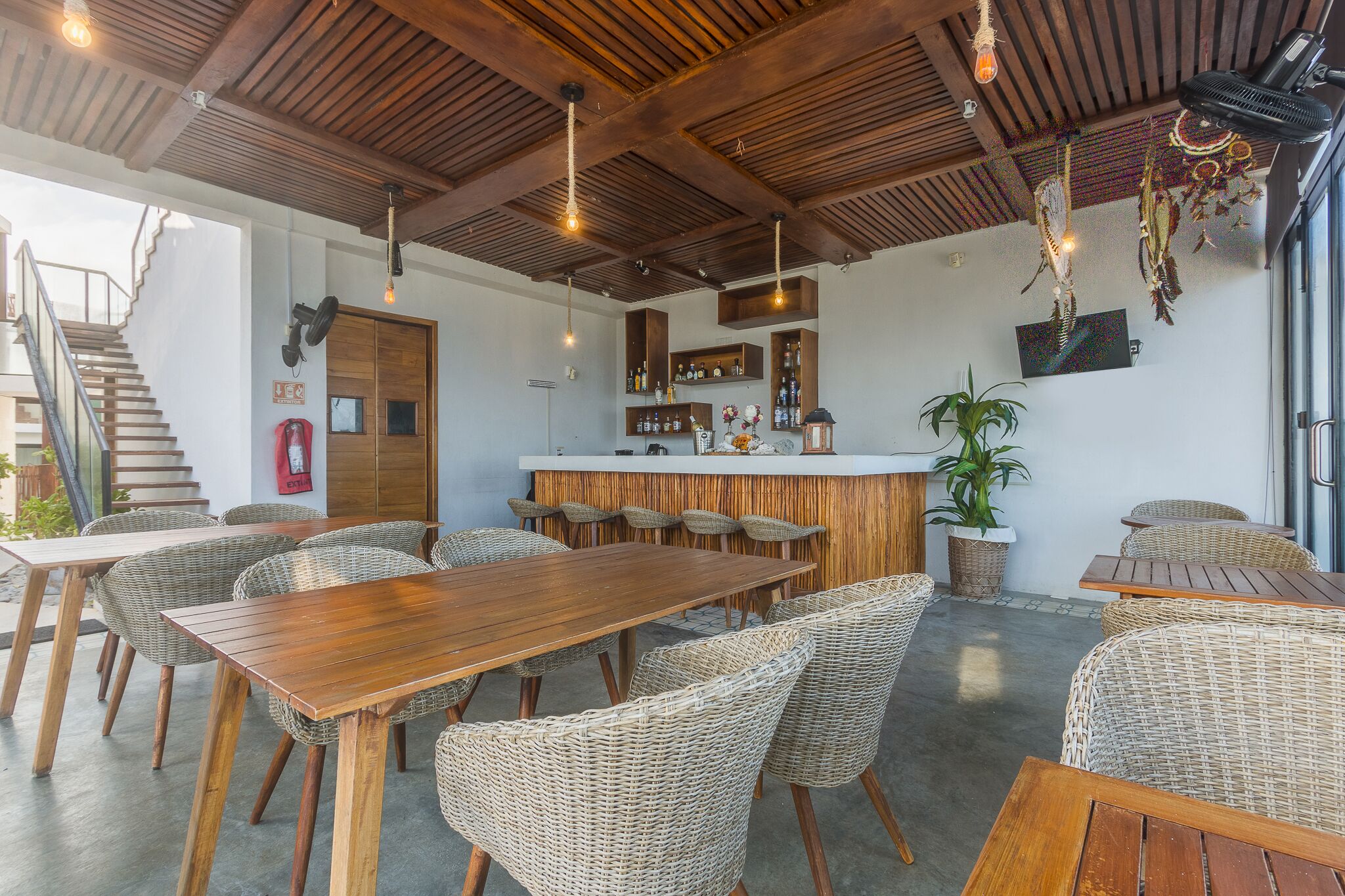 A restaurant with wooden tables and wicker chairs, a bar area, and a staircase leading to an upper level.
