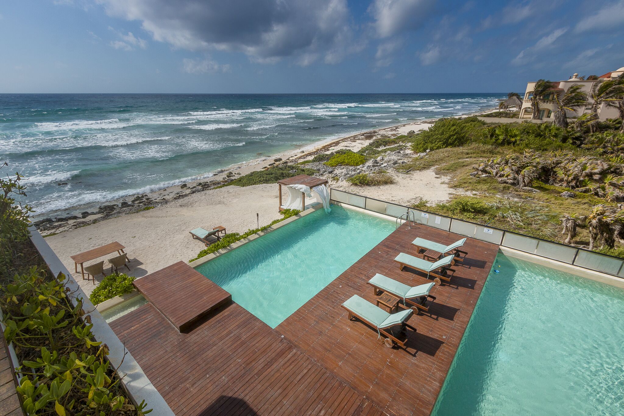 A pool with lounge chairs and a wooden deck overlooking a beach and ocean.