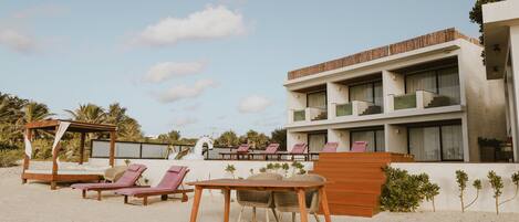 A beachside hotel with a wooden table and lounge chairs, a wooden canopy, and a building with balconies.