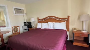 A hotel room with a large bed, a wooden headboard, a bedside table with a lamp, a small desk with a telephone, and an air conditioning unit on the wall.