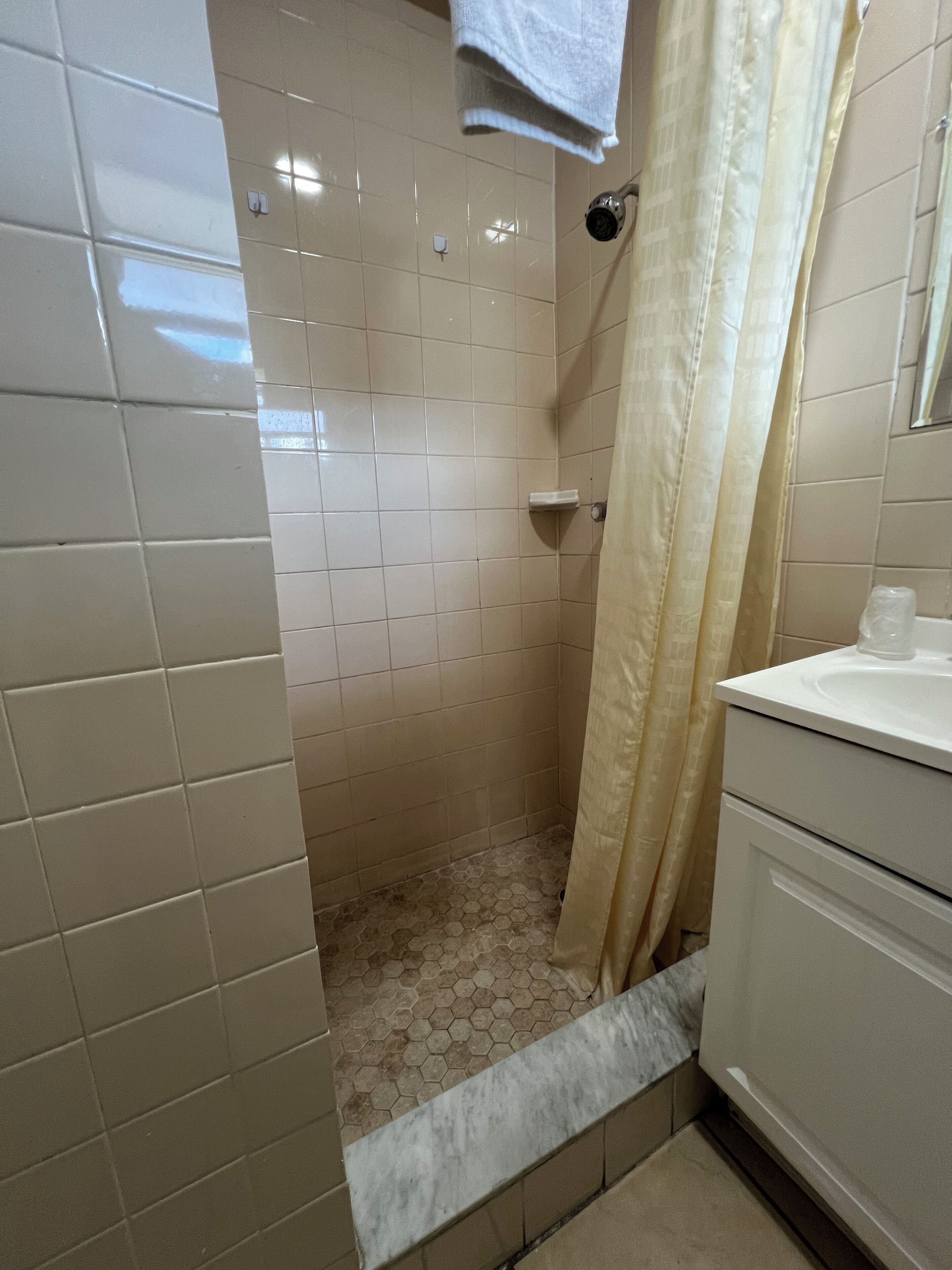 A bathroom with white tiled walls, a shower with a hexagonal floor pattern, a white sink, and a towel hanging on the wall.