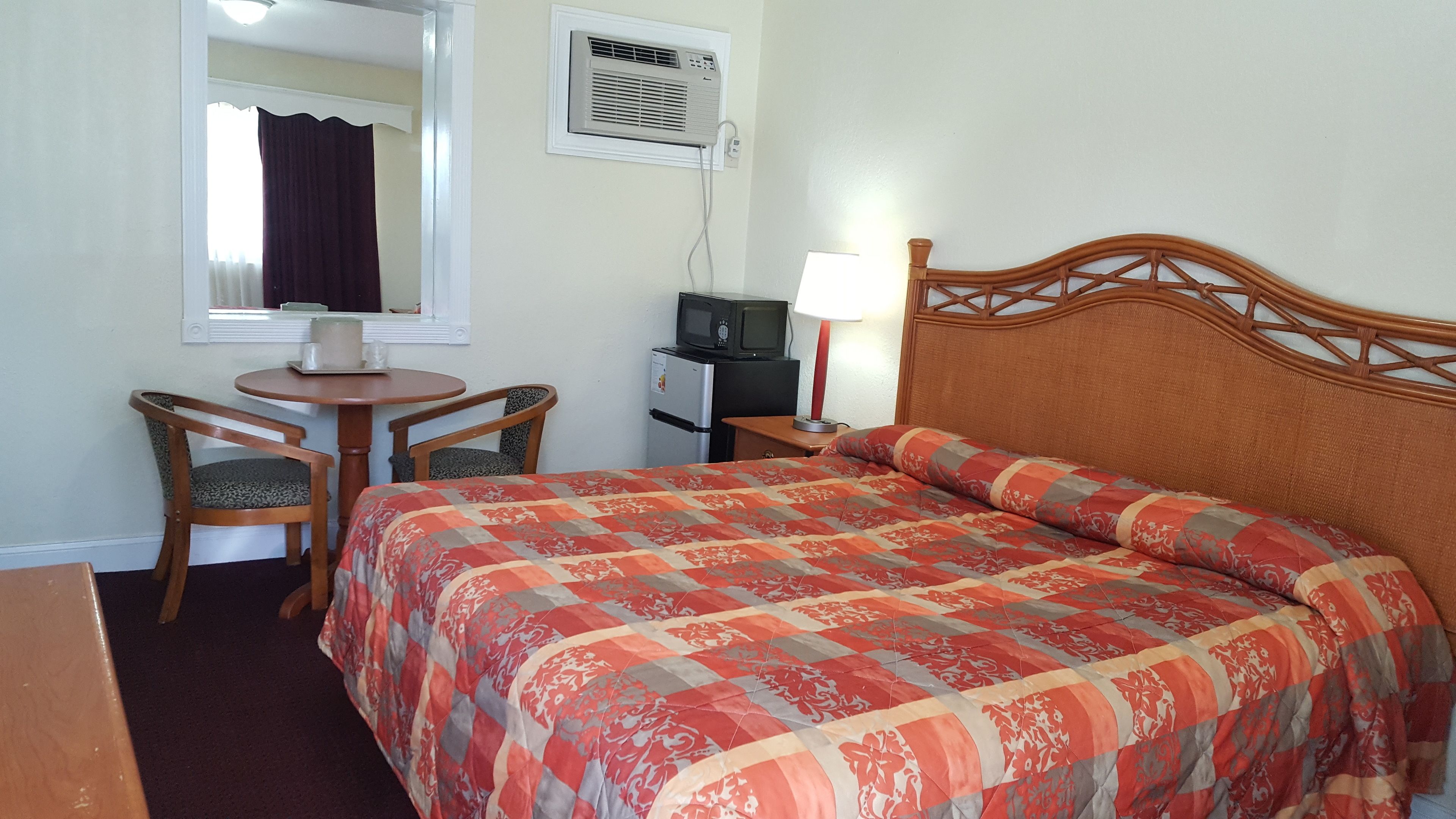 A hotel room with a bed, a desk with a computer, a chair, a lamp, and an air conditioning unit.