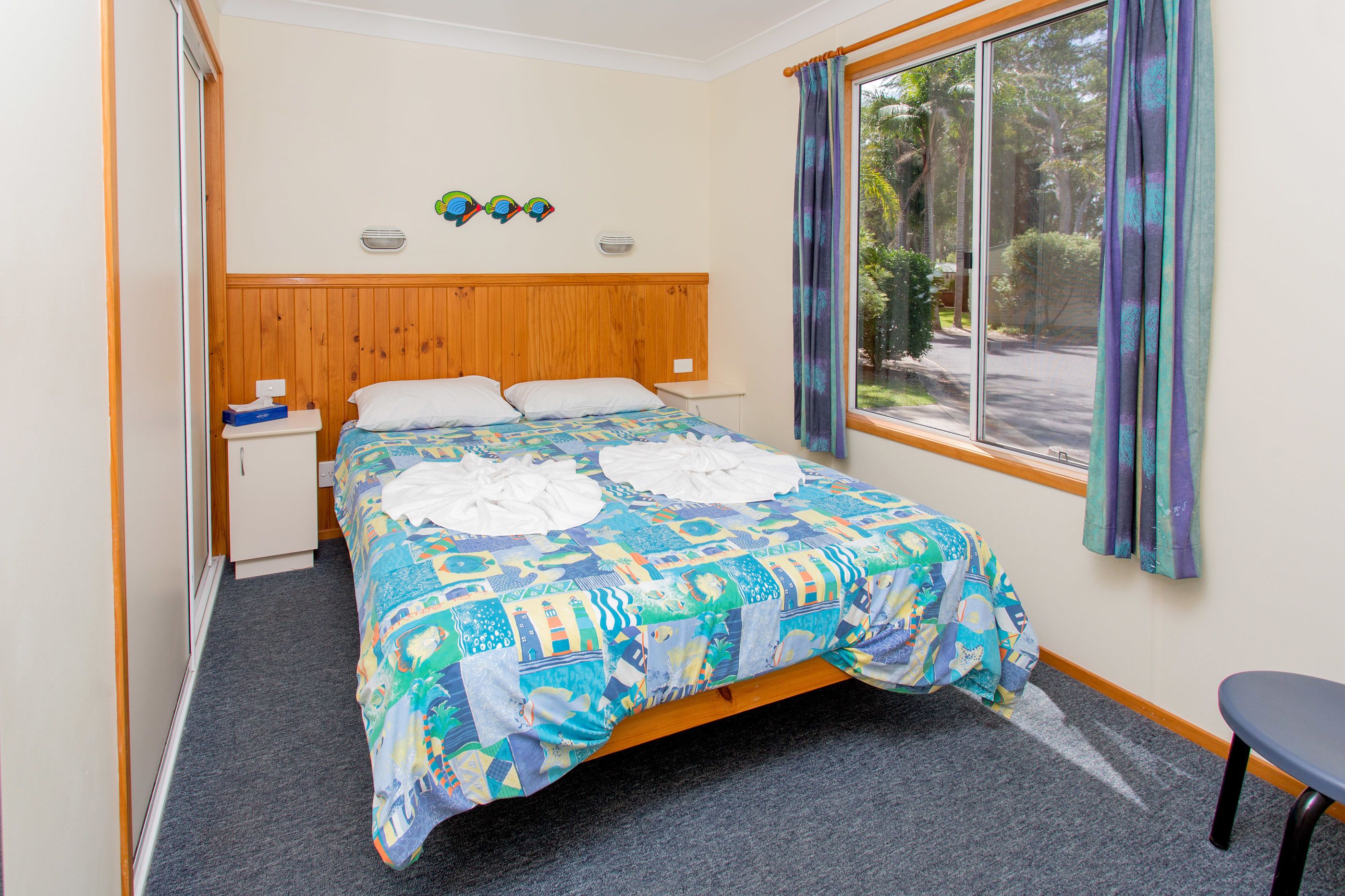 Een slaapkamer met een houten hoofdbord, een bed met een kleurrijke quilt, een klein nachtkastje, een stoel en een raam met gordijnen.