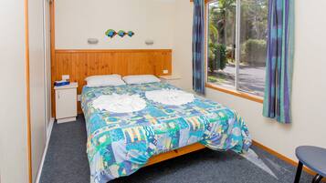 Een slaapkamer met een houten hoofdbord, een bed met een kleurrijke quilt, een klein nachtkastje, een stoel en een raam met gordijnen.