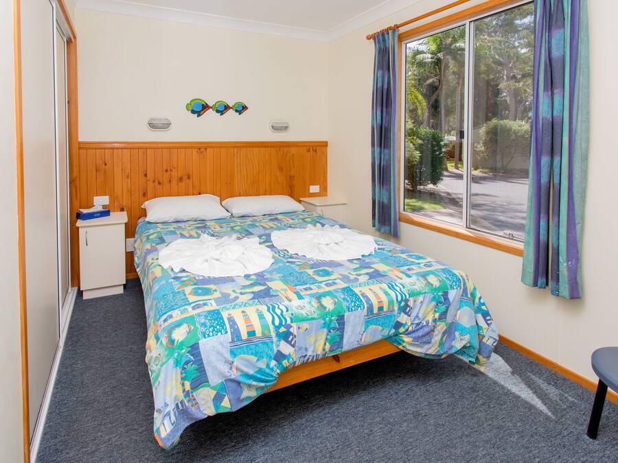 Een slaapkamer met een houten hoofdbord, een bed met een kleurrijke quilt, een klein nachtkastje, een stoel en een raam met gordijnen.