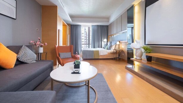 Ein modernes Hotelzimmer mit grauem Sofa, einem runden Couchtisch, einem Bett mit blauer Kopfteil und Blick auf die Stadt durch das Fenster.