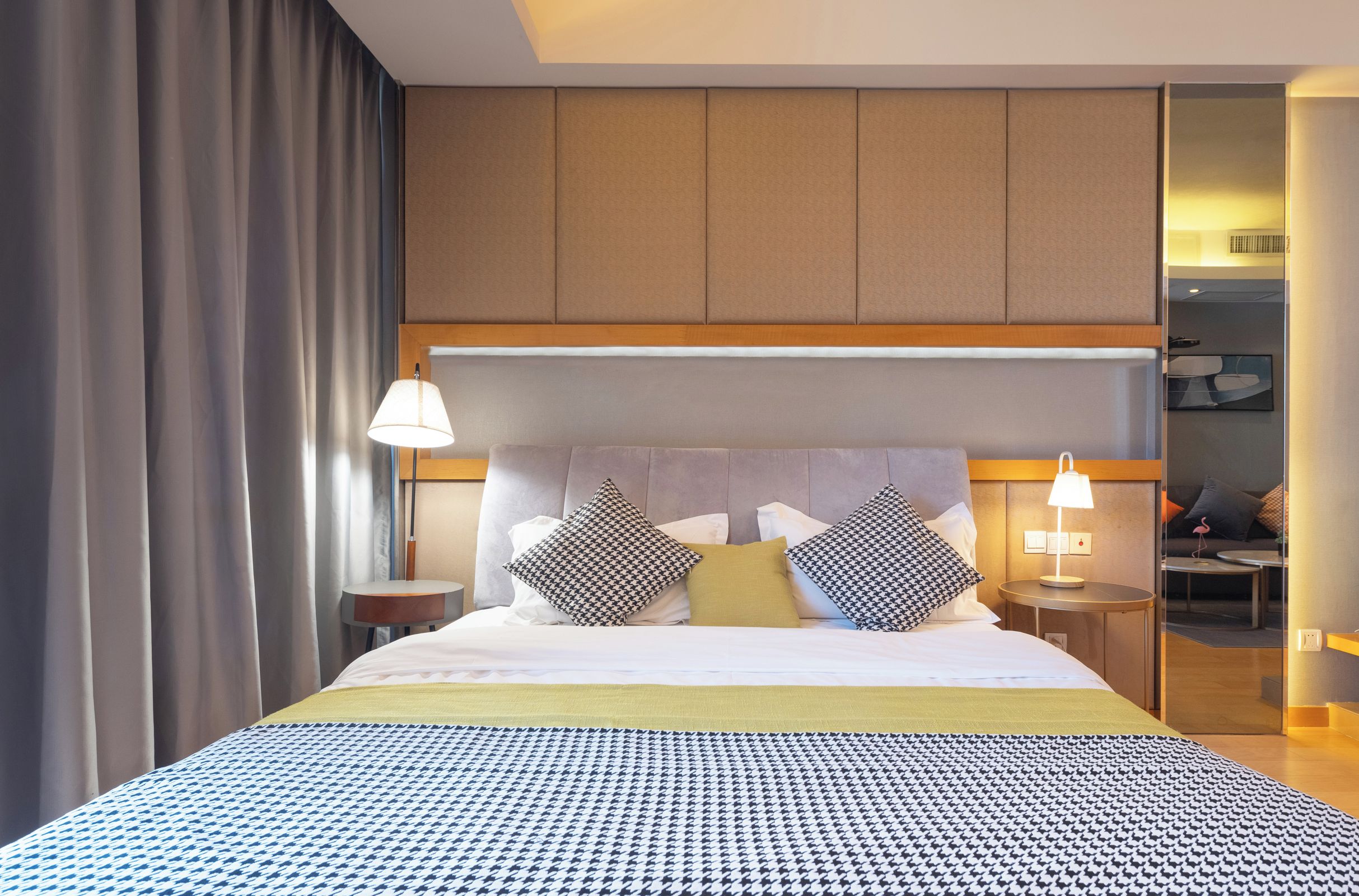 A hotel room with a bed, bedside lamps, a nightstand, and a view of the adjacent room.
