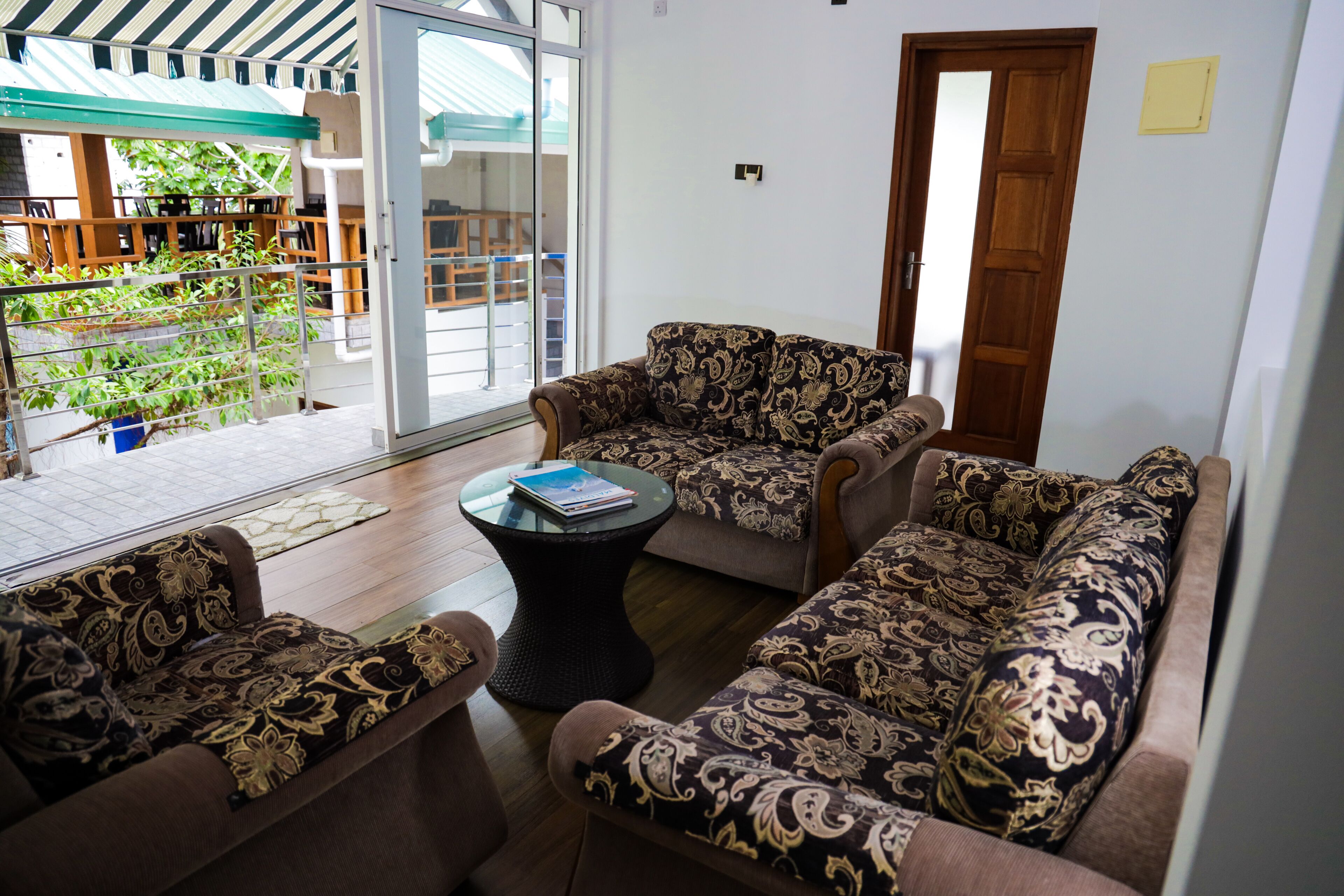 A living area with patterned sofas, a glass coffee table, and a view of a balcony with plants.