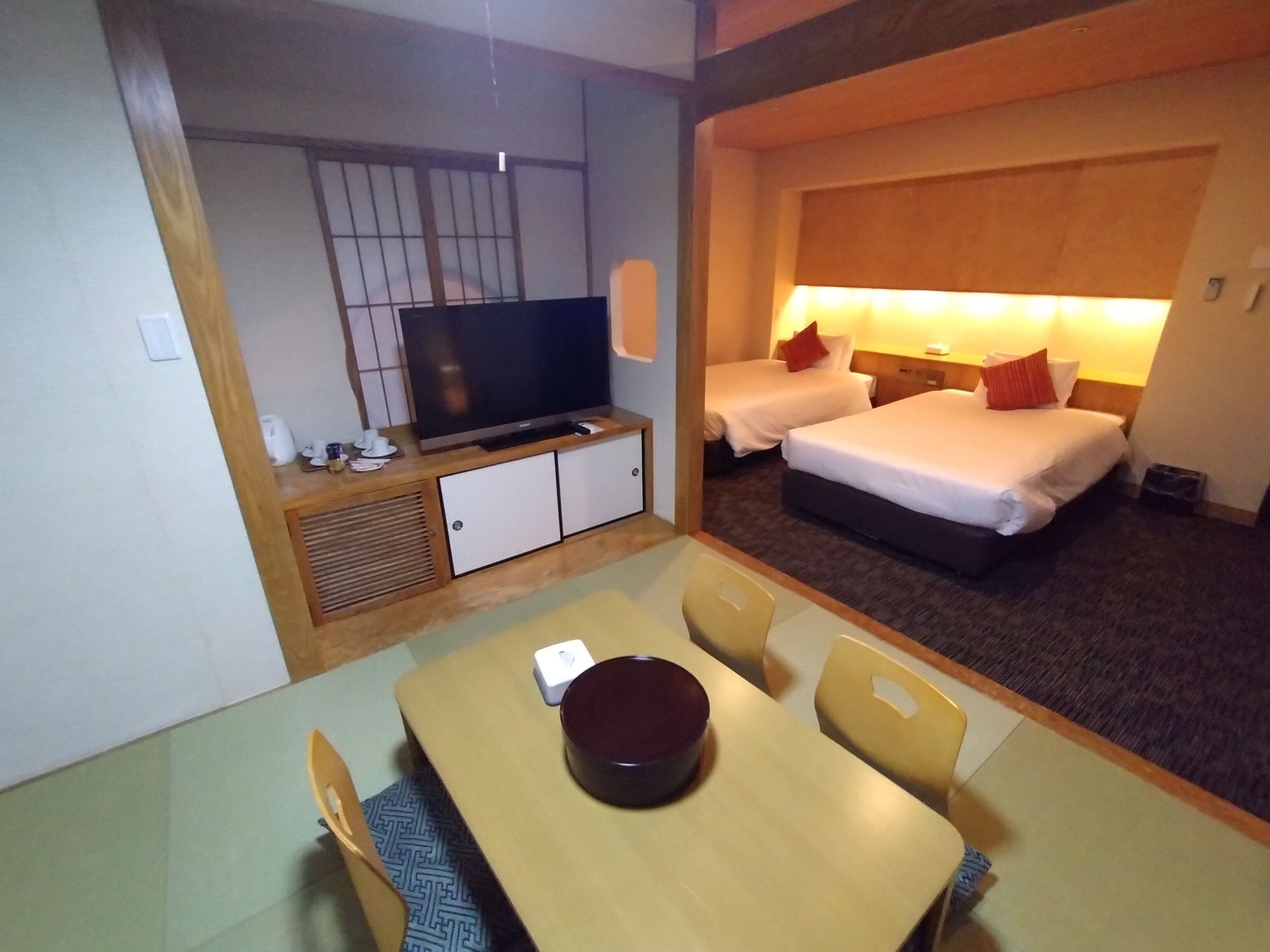 A hotel room with a TV, two beds, a dining area with a table and chairs, and a small kitchenette.