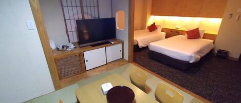 A hotel room with a TV, two beds, a dining area with a table and chairs, and a small kitchenette.