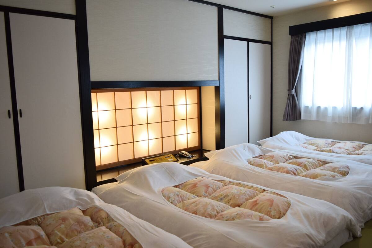 Et hotelværelse med fire senge, en traditionel japansk sengegavl og et stort vindue med gardiner.