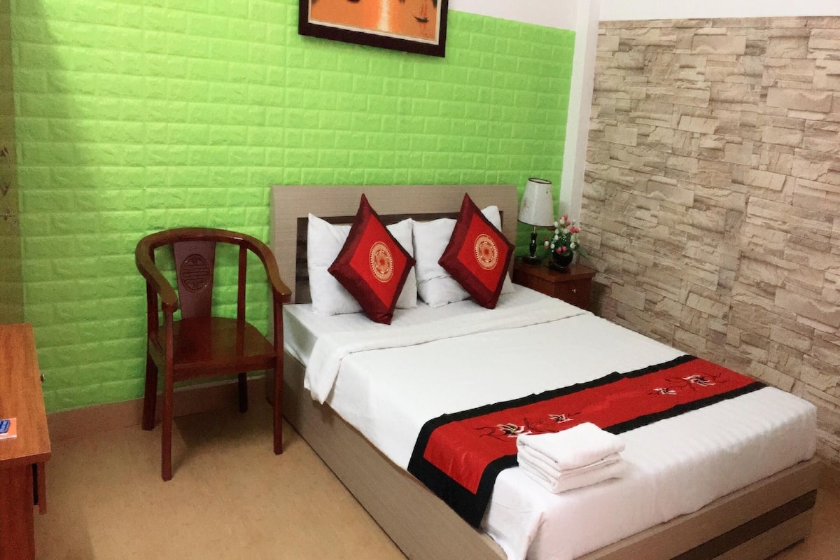 A hotel room with a bed, a chair, a nightstand, and a green brick wall.