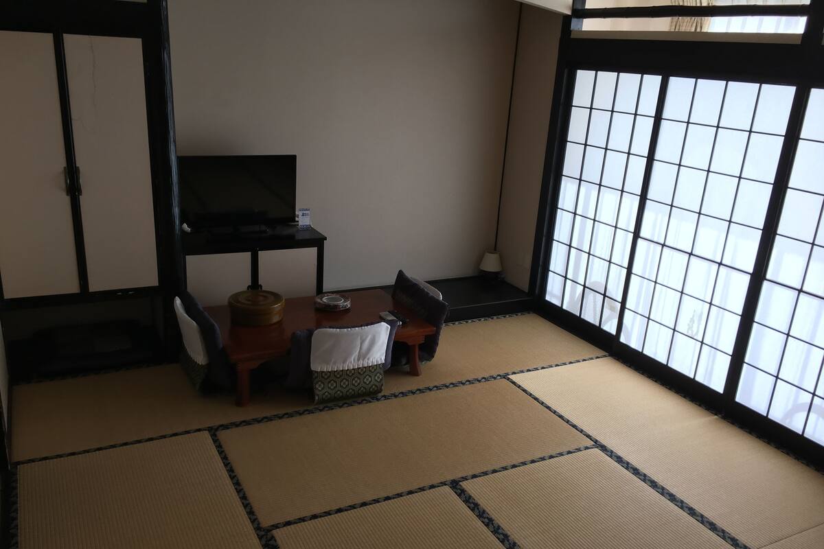A traditional Japanese room with tatami flooring, a low table with a cushion, a television on a stand, and a sliding door with a grid pattern.